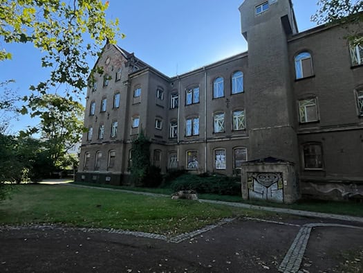 Ein mehrstöckiges, sanierungsbedürftiges historisches Gebäude mit grauem Verputz, Rundbogenfenstern und Graffiti-Verzierungen hinter einer Rasenfläche an der Löbtauer Straße in Dresden.