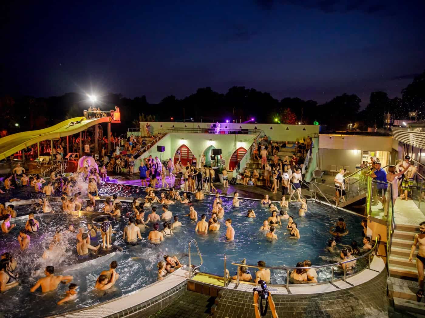 Nächtliche Poolparty in einem beleuchteten Freibad: Zahlreiche Menschen stehen und schwimmen im Wasser, während bunte Lichter die Außenanlage mit einer gelben Wasserrutsche erhellen.