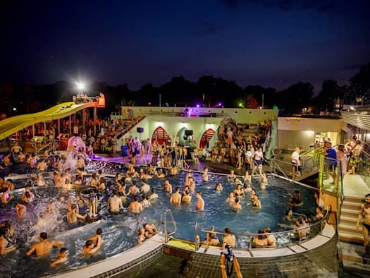 Nächtliche Poolparty in einem beleuchteten Freibad: Zahlreiche Menschen stehen und schwimmen im Wasser, während bunte Lichter die Außenanlage mit einer gelben Wasserrutsche erhellen.
