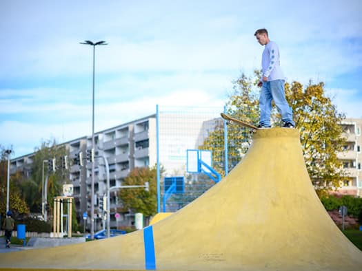 Ein Skater steht mit seinem Board auf der Spitze eines gelben Rampen-Vulkans in der sanierten Streetsport-Anlage am Omsewitzer Ring in Dresden-Gorbitz. Im Hintergrund stehen Wohnblöcke und Bäume.
