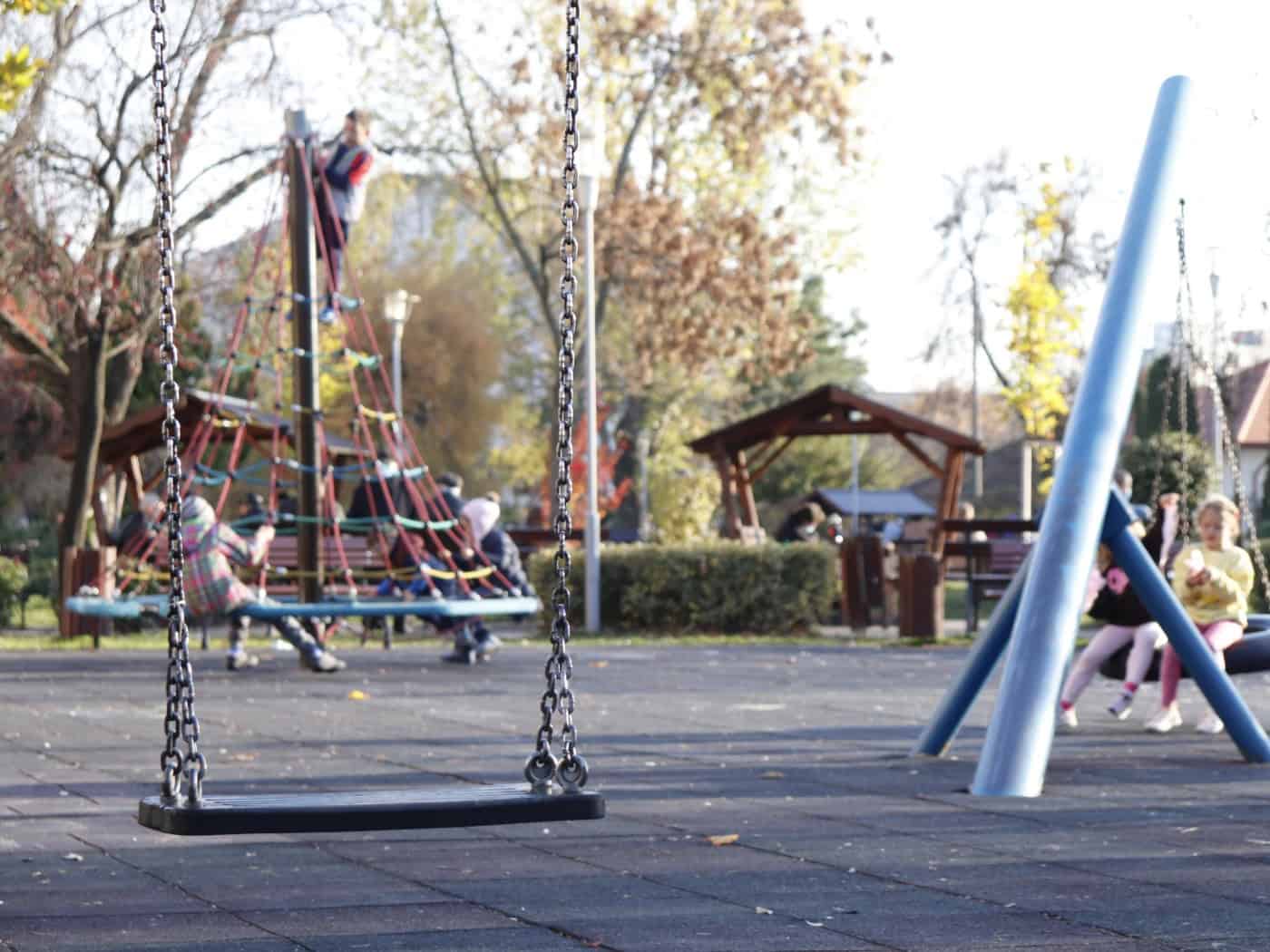 Eine Schaukel an Eisenketten hängt scharf im Vordergrund auf einem Spielplatz. Im leicht unscharfen Hintergrund klettern Kinder auf ein Kletternetz und sitzen auf Spielgeräten in einem sonnigen Park.