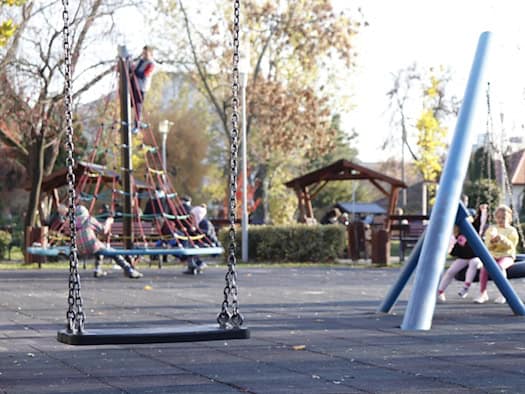 Eine Schaukel an Eisenketten hängt scharf im Vordergrund auf einem Spielplatz. Im leicht unscharfen Hintergrund klettern Kinder auf ein Kletternetz und sitzen auf Spielgeräten in einem sonnigen Park.
