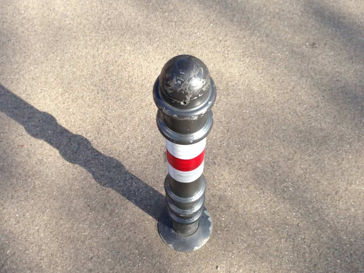 Ein grauer Metallpoller mit rot-weißer Markierung steht auf einer gepflasterten Fläche und wirft einen langen Schatten. Die Nahaufnahme zeigt die zylinderförmige Absperrung von oben.