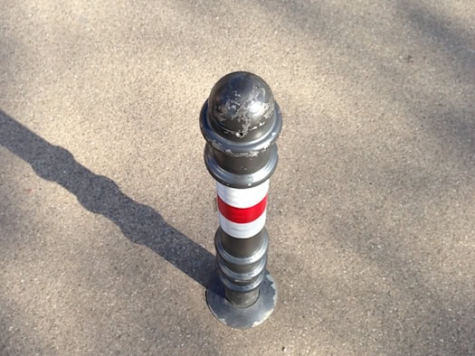 Ein grauer Metallpoller mit rot-weißer Markierung steht auf einer gepflasterten Fläche und wirft einen langen Schatten. Die Nahaufnahme zeigt die zylinderförmige Absperrung von oben.