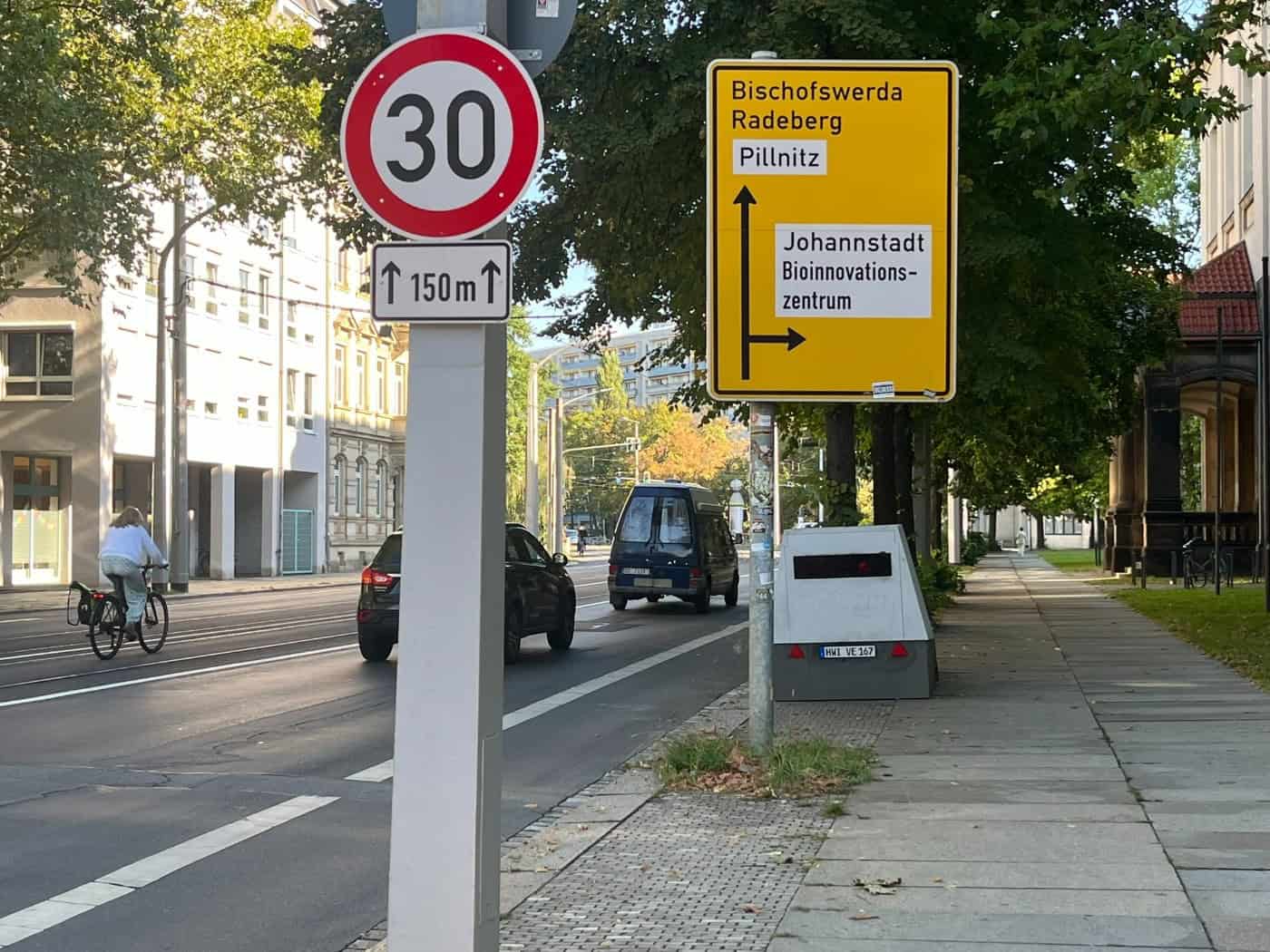 Ein grauer Anhänger-Superblitzer steht auf dem Gehweg der Güntzstraße hinter einem Tempo-30-Schild und einem gelben Wegweiser. Auf der asphaltierten Straße fahren Autos und ein Fahrrad.