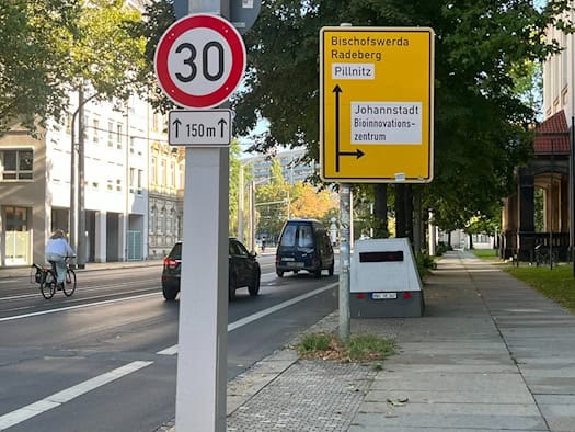 Ein grauer Anhänger-Superblitzer steht auf dem Gehweg der Güntzstraße hinter einem Tempo-30-Schild und einem gelben Wegweiser. Auf der asphaltierten Straße fahren Autos und ein Fahrrad.