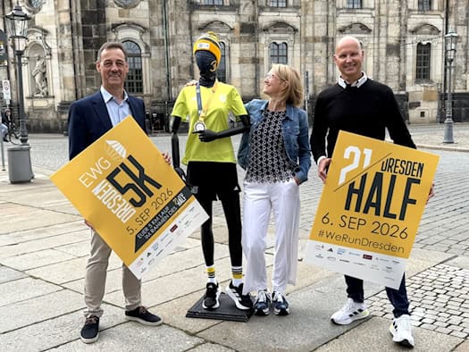 Michael Reichel, Antje Neelmeijer und André Egger halten Plakate für den DresdenHALF und EWG Dresden5K. Zwischen ihnen steht eine Schaufensterpuppe in Laufbekleidung vor Altstadtkulisse.