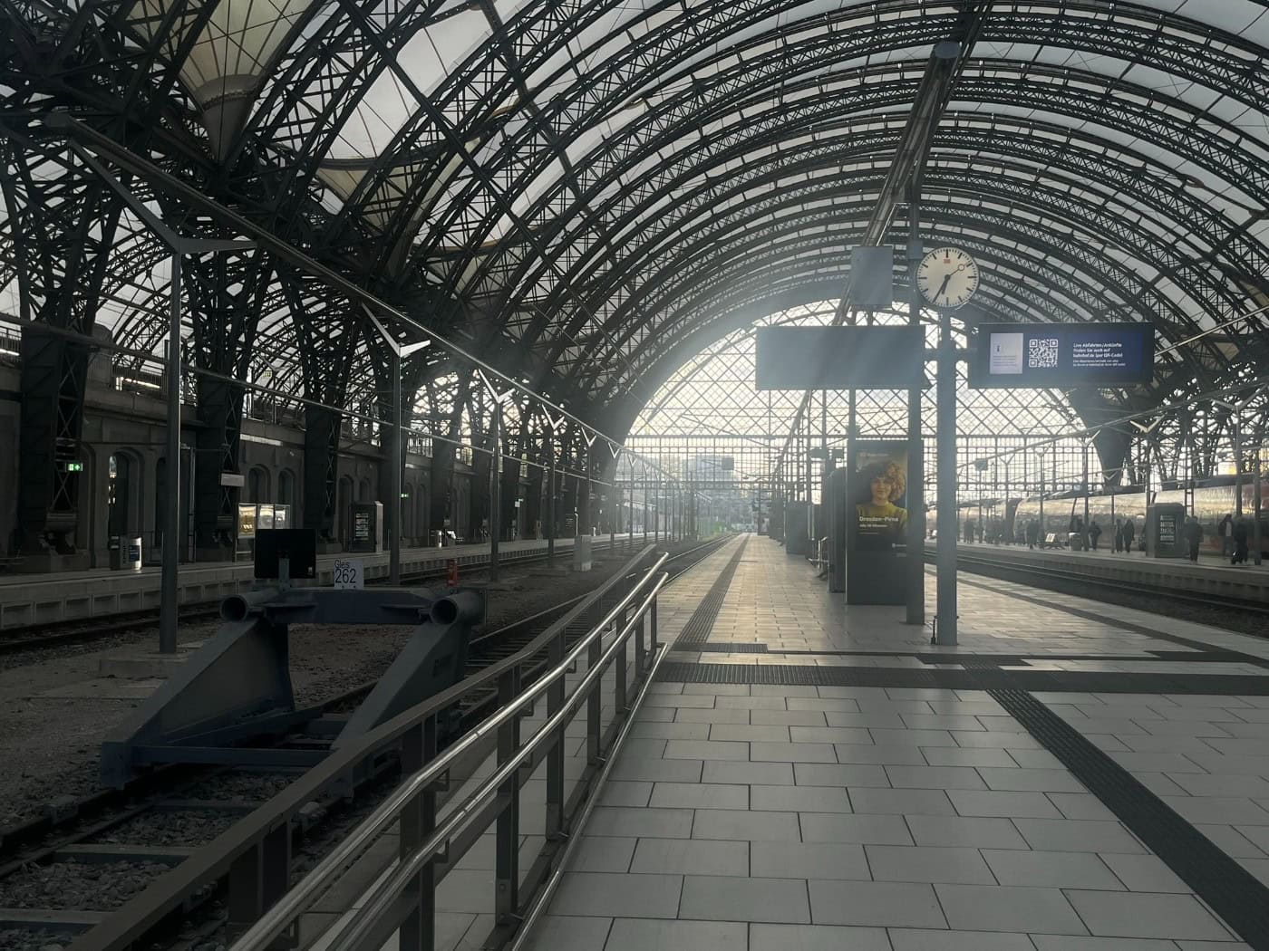 Blick durch die große, von einer Stahlkonstruktion überspannte Halle des Dresdner Hauptbahnhofs. Links liegen Gleise mit einem Prellbock, rechts ein Bahnsteig mit Bahnhofsuhr, Anzeigetafeln und einem wartenden Zug im hellen Tageslicht.