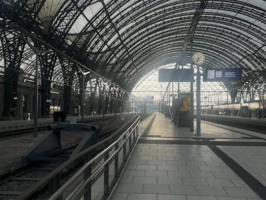 Blick durch die große, von einer Stahlkonstruktion überspannte Halle des Dresdner Hauptbahnhofs. Links liegen Gleise mit einem Prellbock, rechts ein Bahnsteig mit Bahnhofsuhr, Anzeigetafeln und einem wartenden Zug im hellen Tageslicht.