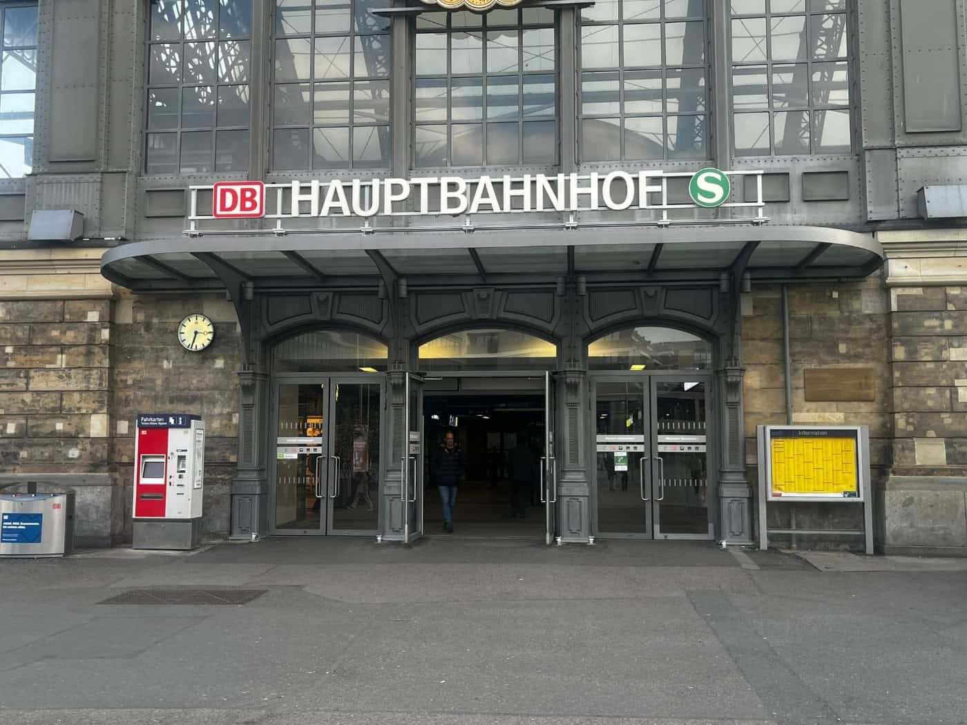 Haupteingang des Dresdner Hauptbahnhofs mit historischem Steinportal, großer Glasfront, DB-Logo, Fahrscheinautomat und Bahnhofsuhr. Eine Person geht durch die gläserne Eingangstür.