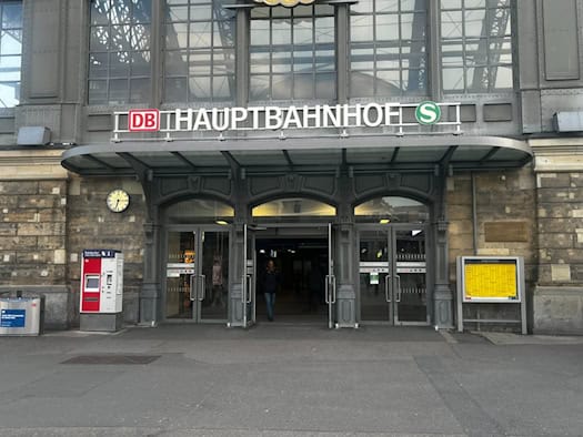 Haupteingang des Dresdner Hauptbahnhofs mit historischem Steinportal, großer Glasfront, DB-Logo, Fahrscheinautomat und Bahnhofsuhr. Eine Person geht durch die gläserne Eingangstür.