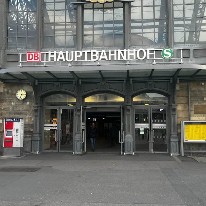 Haupteingang des Dresdner Hauptbahnhofs mit historischem Steinportal, großer Glasfront, DB-Logo, Fahrscheinautomat und Bahnhofsuhr. Eine Person geht durch die gläserne Eingangstür.