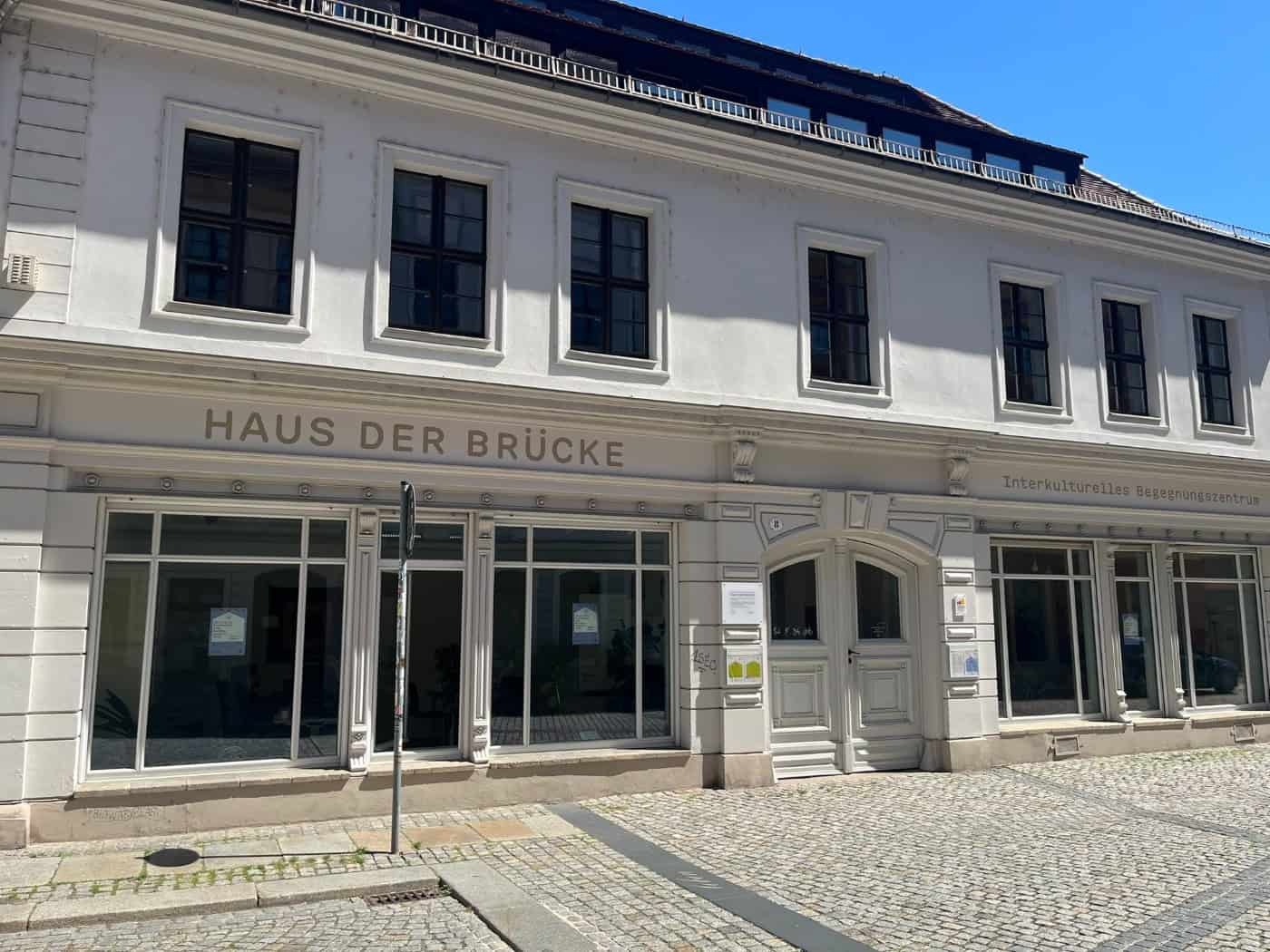 Außenansicht des zweigeschossigen, weiß gestrichenen Gebäudes „Haus der Brücke“ in der Dresdner Rähnitzgasse mit großflächigen Schaufenstern, historischer Holztür und gepflasterter Straße bei sonnigem Wetter.