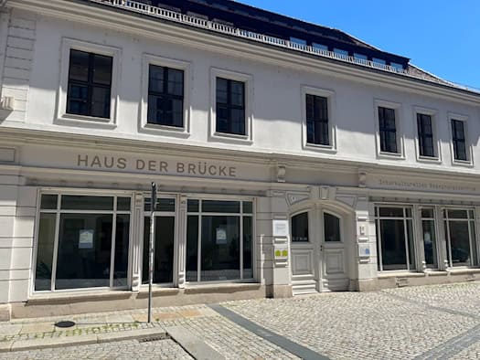 Außenansicht des zweigeschossigen, weiß gestrichenen Gebäudes „Haus der Brücke“ in der Dresdner Rähnitzgasse mit großflächigen Schaufenstern, historischer Holztür und gepflasterter Straße bei sonnigem Wetter.