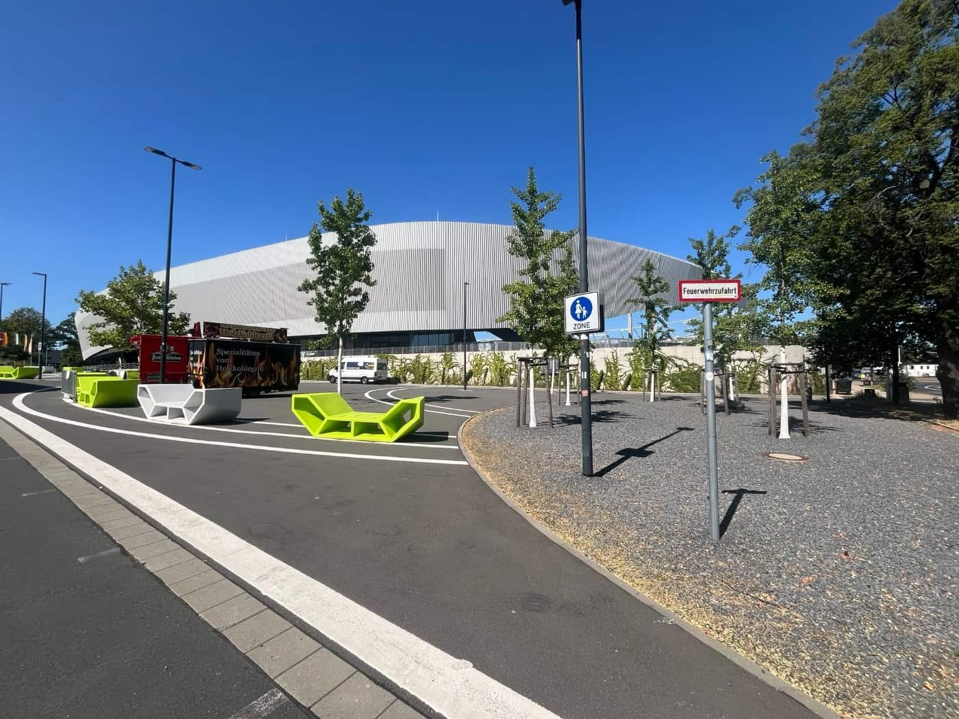 Blick über den asphaltierten Vorplatz des modernen Heinz-Steyer-Stadions in Dresden mit auffälligen leuchtgrünen Sitzbänken, jungen Bäumen und Verkehrsschildern unter hellem blauen Himmel.