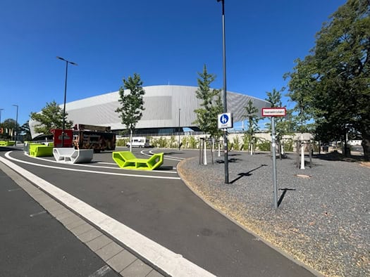 Blick über den asphaltierten Vorplatz des modernen Heinz-Steyer-Stadions in Dresden mit auffälligen leuchtgrünen Sitzbänken, jungen Bäumen und Verkehrsschildern unter hellem blauen Himmel.