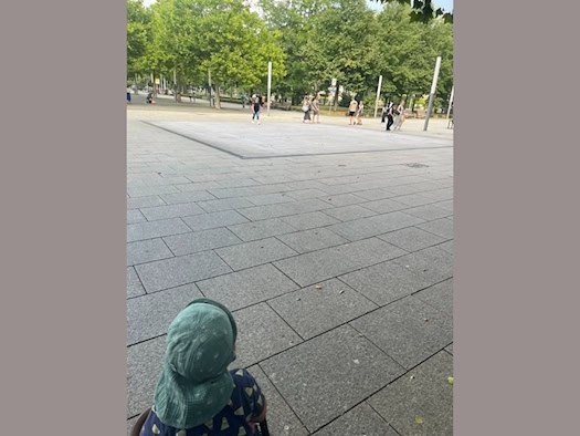 Ein Kind mit grüner Mütze im Kinderwagen blickt auf einen großen, gepflasterten Platz (Jorge-Gomondai-Platz, Dresden). Im Mittelgrund die trockene Granitfläche des Bodenbrunnens. Mehrere Passanten gehen am Rand entlang. Grüne Bäume und Lichtstelen säumen den Platz.