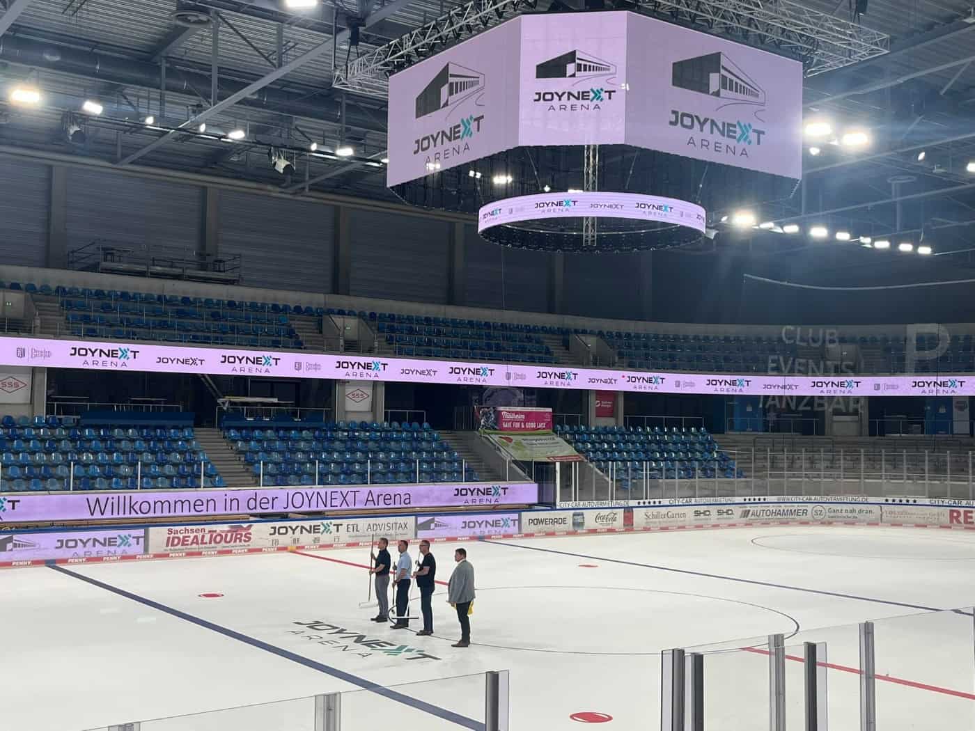 Blick in die große, leere Eishalle der JOYNEXT Arena Dresden mit glatter Eisfläche, auf der vier Personen stehen, umgeben von leeren Zuschauertribünen und dem leuchtenden Würfel-Display an der Decke.