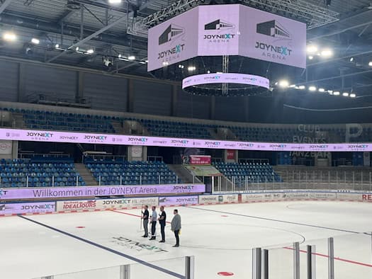 Blick in die große, leere Eishalle der JOYNEXT Arena Dresden mit glatter Eisfläche, auf der vier Personen stehen, umgeben von leeren Zuschauertribünen und dem leuchtenden Würfel-Display an der Decke.