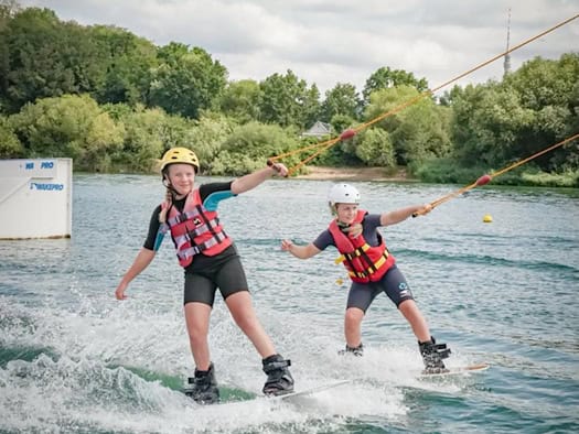 Zwei Mädchen in Neoprenanzügen, Schwimmwesten und Helmen fahren nebeneinander auf Wakeboards über das Wasser des Kiessees Leuben. Sie halten sich an Zugseilen fest und erzeugen Gischt. Im Hintergrund stehen grüne Bäume.