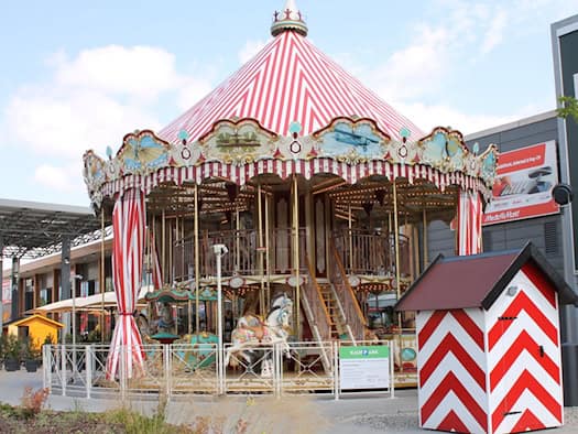 Zweistöckiges Nostalgie-Karussell mit rot-weiß gestreiftem Dach und geschnitzten Pferden auf einem Platz vor einem Einkaufszentrum. Daneben steht ein rot-weiß gestreiftes Kassenhäuschen im Sonnenlicht.