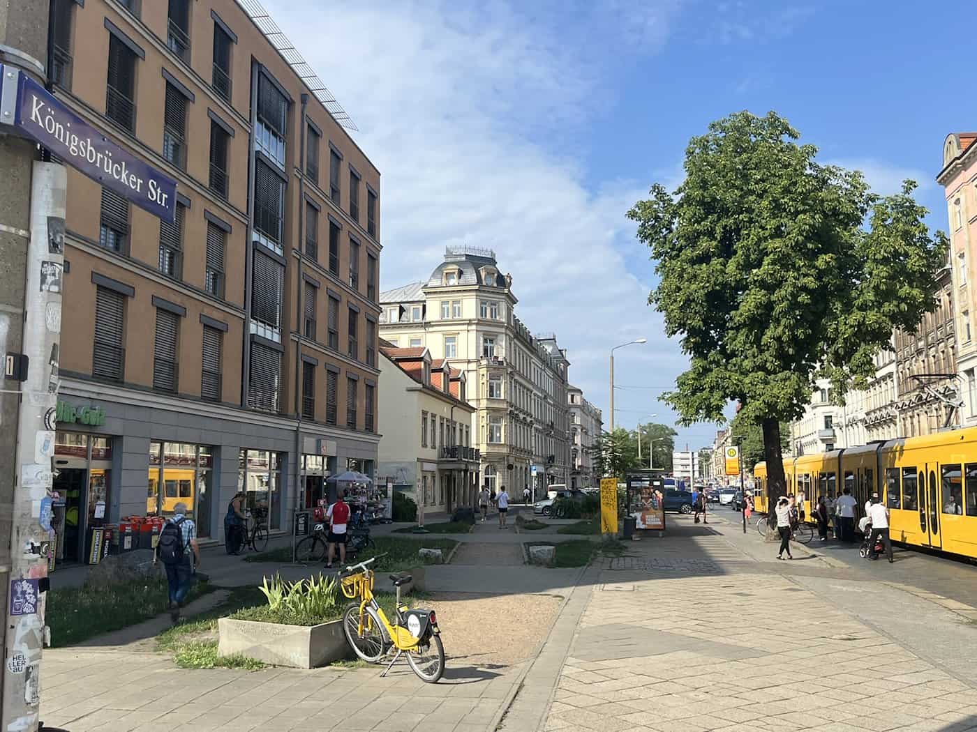 Blick auf die Königsbrücker Straße in Dresden mit einem Straßenschild im Vordergrund. Auf dem breiten Gehweg parkt ein gelbes Leihfahrrad, rechts steht eine gelbe Straßenbahn der DVB an einer Haltestelle vor Gründerzeitgebäuden und Bäumen.