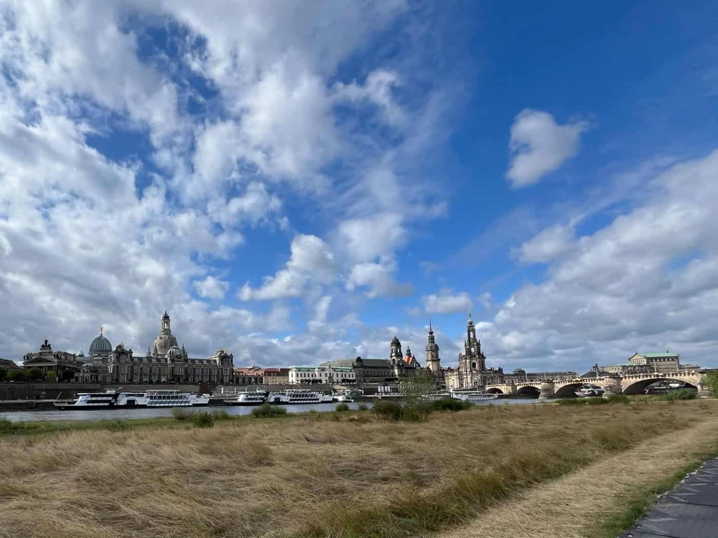 Panorama der Dresdner Altstadt mit Frauenkirche und Hofkirche, gesehen von den trockenen Elbwiesen unter leicht bewölktem blauem Himmel. Am Flussufer liegen Passagierschiffe vor Anker.