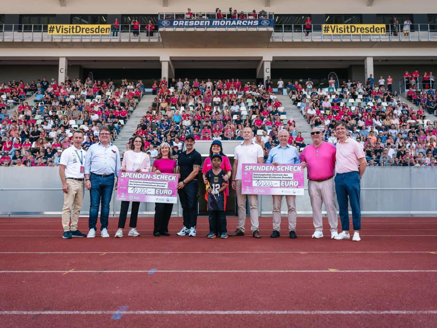 Vertreter der Dresden Monarchs und medizinischer Zentren stehen auf der Laufbahn des Heinz-Steyer-Stadions und halten zwei pinke Spendenschecks über jeweils 10.000 Euro. Im Hintergrund sitzt das Publikum auf der gefüllten Tribüne.