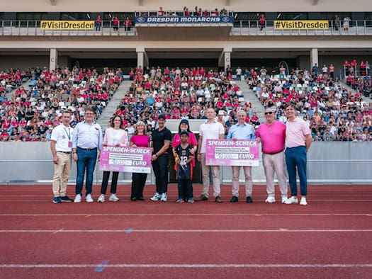 Vertreter der Dresden Monarchs und medizinischer Zentren stehen auf der Laufbahn des Heinz-Steyer-Stadions und halten zwei pinke Spendenschecks über jeweils 10.000 Euro. Im Hintergrund sitzt das Publikum auf der gefüllten Tribüne.
