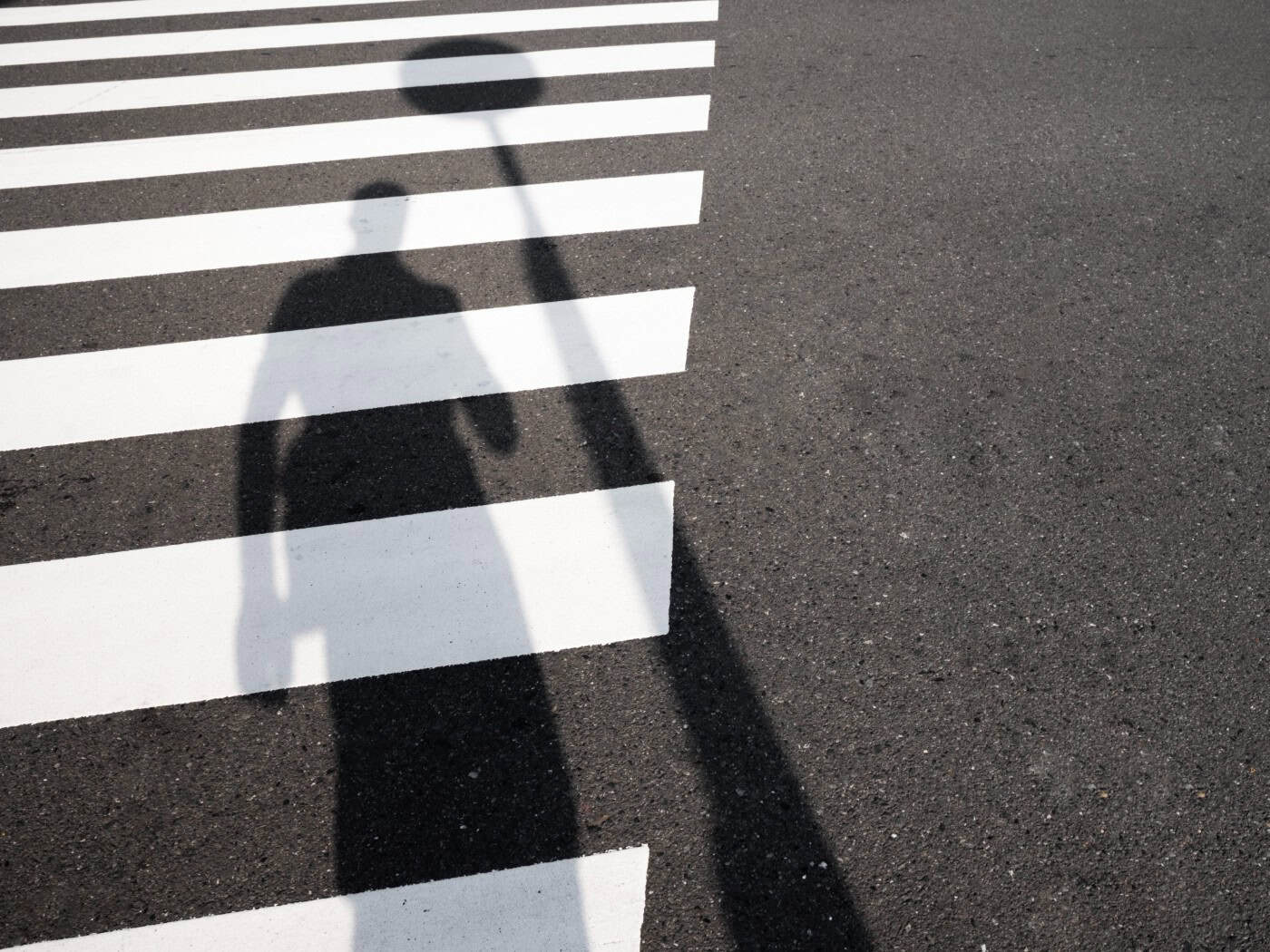 Schatten einer Person fällt auf die weißen Streifen eines Zebrastreifens auf dunklem Asphalt. (Symbolbild zur Verkehrssicherheit und Fußgängerüberwegen)