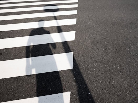 Schatten einer Person fällt auf die weißen Streifen eines Zebrastreifens auf dunklem Asphalt. (Symbolbild zur Verkehrssicherheit und Fußgängerüberwegen)