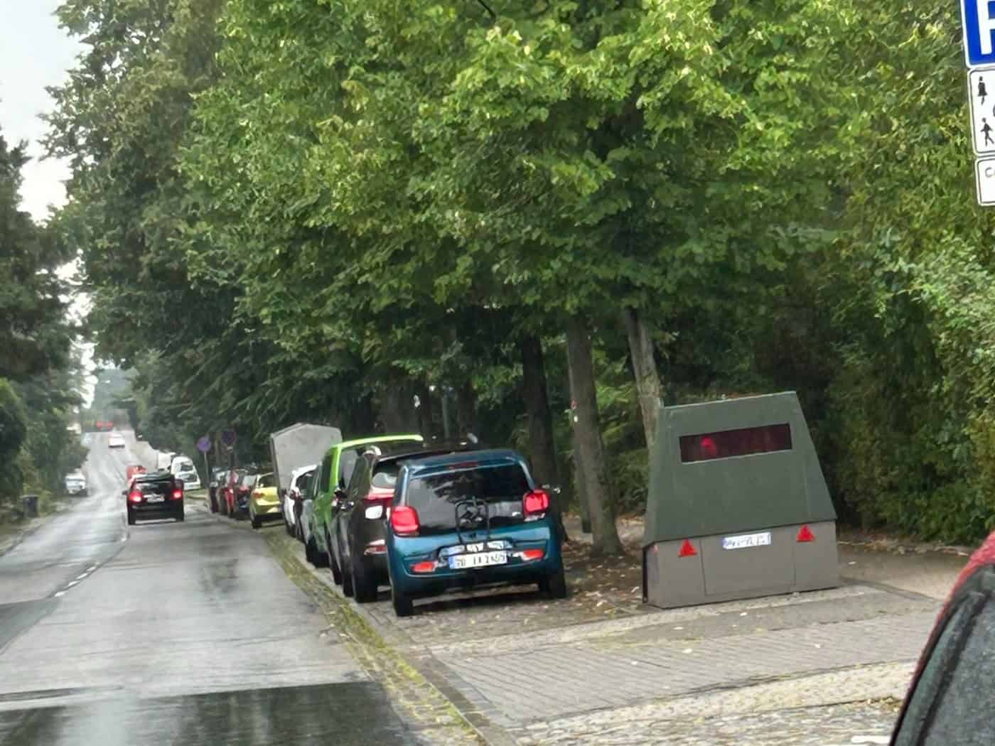 Ein olivgrüner Blitzer-Anhänger steht am Rand einer nassen Straße unter Bäumen neben geparkten Autos an der Pillnitzer Landstraße in Dresden.