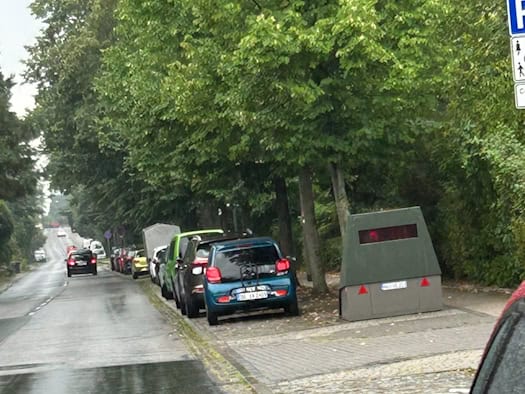 Ein olivgrüner Blitzer-Anhänger steht am Rand einer nassen Straße unter Bäumen neben geparkten Autos an der Pillnitzer Landstraße in Dresden.