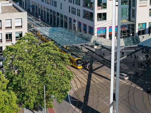 Luftaufnahme der DVB-Haltestelle am Dresdner Postplatz: Eine gelbe Straßenbahn steht unter dem modernen Glasdach der Haltestellenüberdachung. Daneben erstreckt sich ein großer grüner Baum auf dem Platz.
