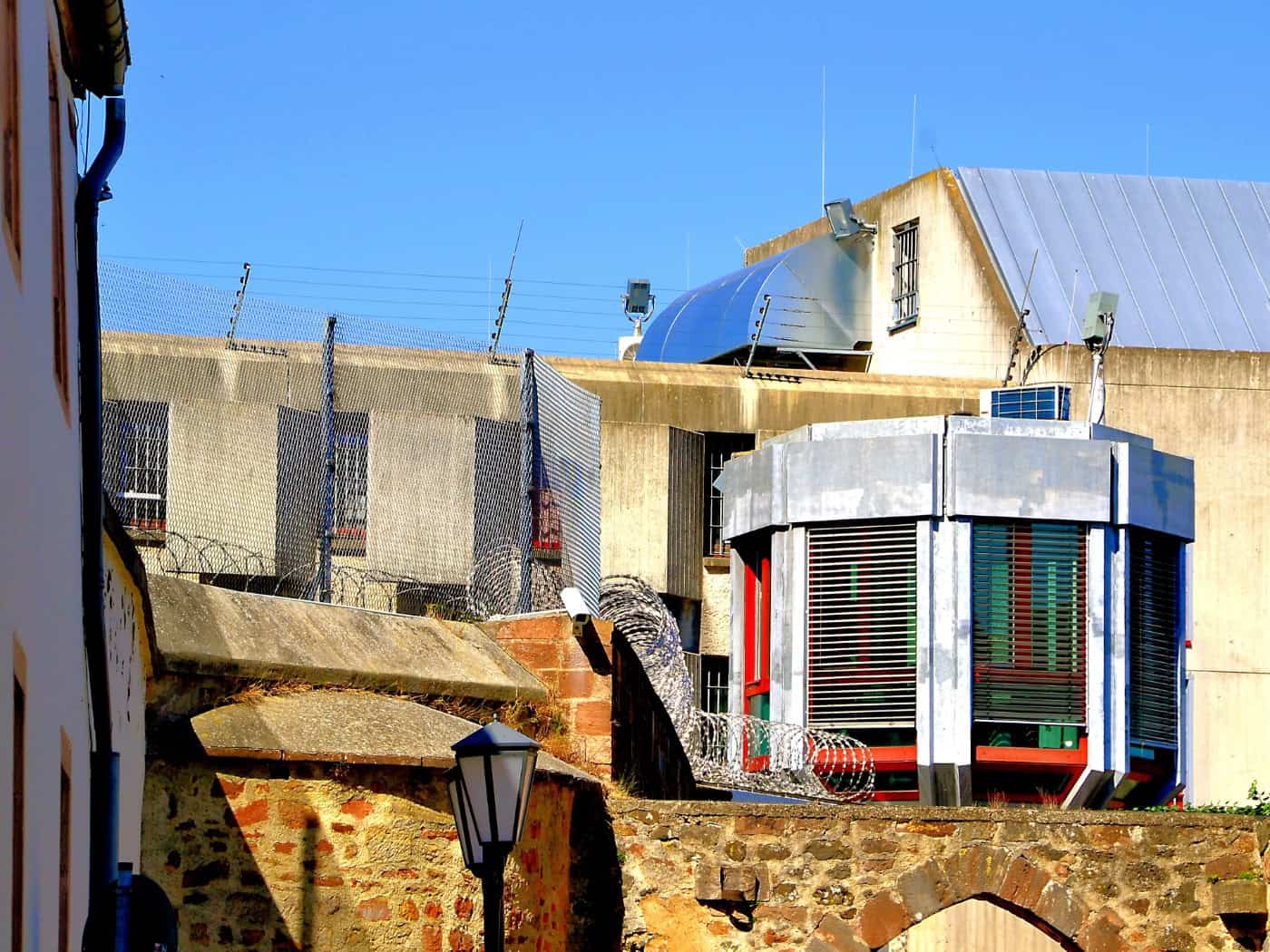 Außenansicht einer Gefängnisanlage mit hohen Steinmauern, Stacheldrahtzäunen und einem modernen Sicherheitsgebäude vor Betonbauten unter blauem Himmel.