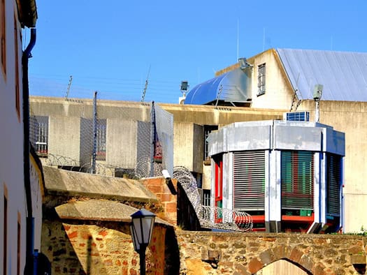 Außenansicht einer Gefängnisanlage mit hohen Steinmauern, Stacheldrahtzäunen und einem modernen Sicherheitsgebäude vor Betonbauten unter blauem Himmel.