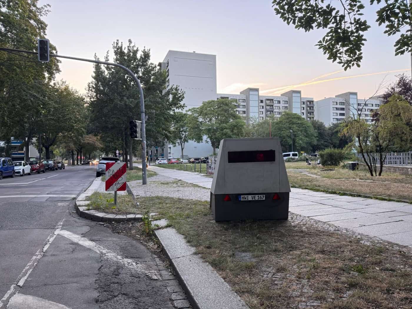 Ein grauer teilstationärer Blitzer-Anhänger mit dem Kennzeichen HWI-VE 167 steht am Rand der Reichenbachstraße in Dresden. Im Hintergrund sind Fahrbahn, Ampel und Plattenbauten zu sehen.