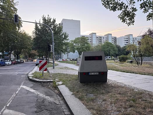 Ein grauer teilstationärer Blitzer-Anhänger mit dem Kennzeichen HWI-VE 167 steht am Rand der Reichenbachstraße in Dresden. Im Hintergrund sind Fahrbahn, Ampel und Plattenbauten zu sehen.