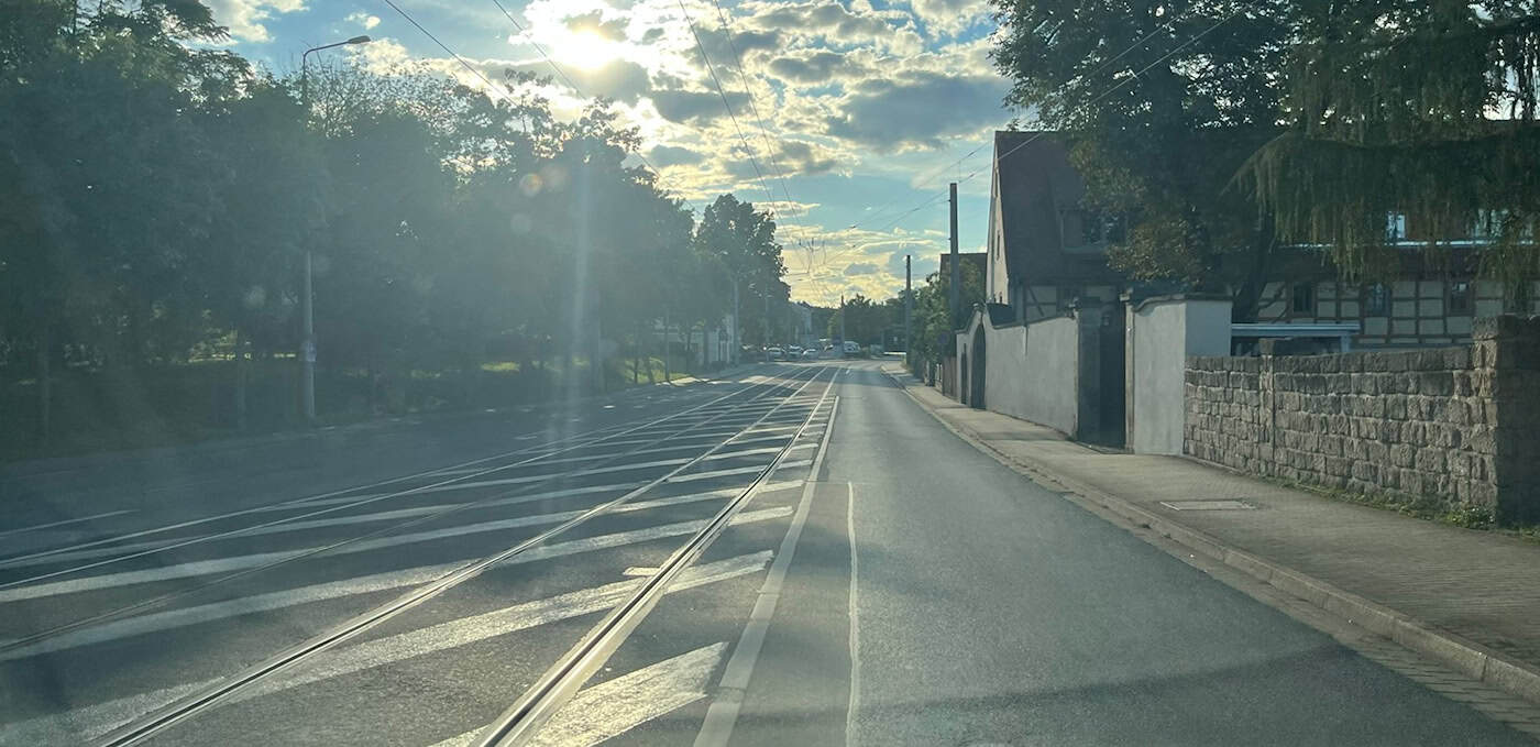 Perspektive aus der Windschutzscheibe eines Autos auf die Mügelner Straße in Dresden-Reick mit Straßenbahngleisen. Die Sonne steht tief über der asphaltierten Fahrbahn in Richtung Altreick.