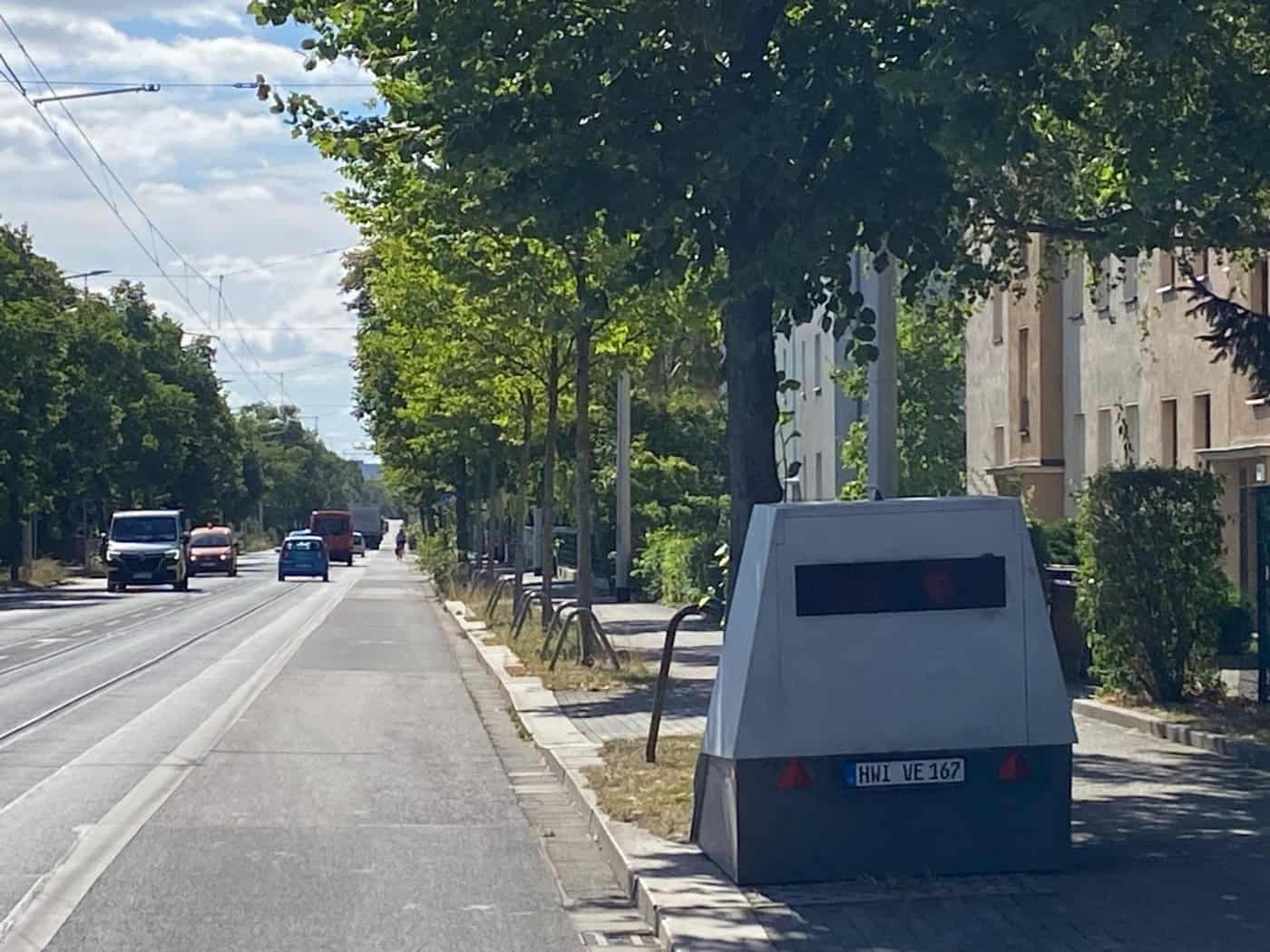 Grauer mobiler Messanhänger mit Kennzeichen HWI VE 167 steht auf dem Gehweg unter Bäumen an einer Straße in Dresden. Auf der mehrspurigen Fahrbahn daneben rollt der Verkehr.