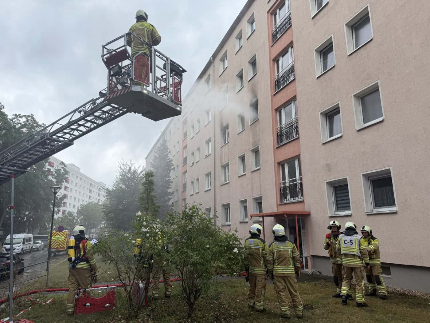 Einsatzkräfte der Feuerwehr löschen von einer Drehleiter aus einen Wohnungsbrand an einem Mehrfamilienhaus in Dresden. Am Boden stehen mehrere Feuerwehrmänner in Schutzkleidung.
