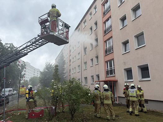 Einsatzkräfte der Feuerwehr löschen von einer Drehleiter aus einen Wohnungsbrand an einem Mehrfamilienhaus in Dresden. Am Boden stehen mehrere Feuerwehrmänner in Schutzkleidung.