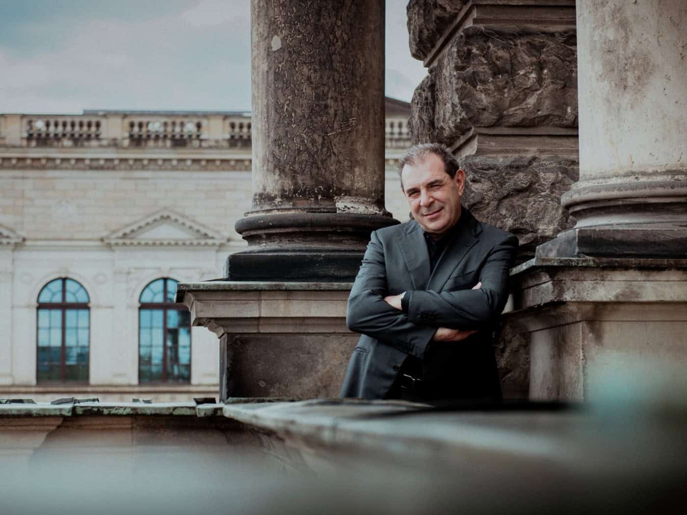 Chefdirigent Daniele Gatti lehnt im dunklen Sakko mit verschränkten Armen an Sandsteinsäulen der Dresdner Semperoper und blickt lächelnd in die Kamera.