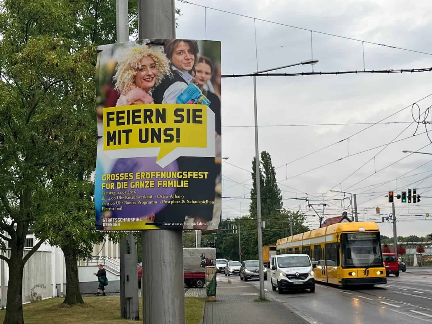 Ein Plakat am Straßenrand wirbt für das Eröffnungsfest des Staatsschauspiels Dresden am 22. August 2026. Im Hintergrund fährt eine gelbe Dresdner Straßenbahn an einer Straße entlang.
