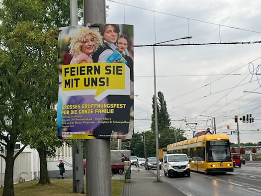Ein Plakat am Straßenrand wirbt für das Eröffnungsfest des Staatsschauspiels Dresden am 22. August 2026. Im Hintergrund fährt eine gelbe Dresdner Straßenbahn an einer Straße entlang.