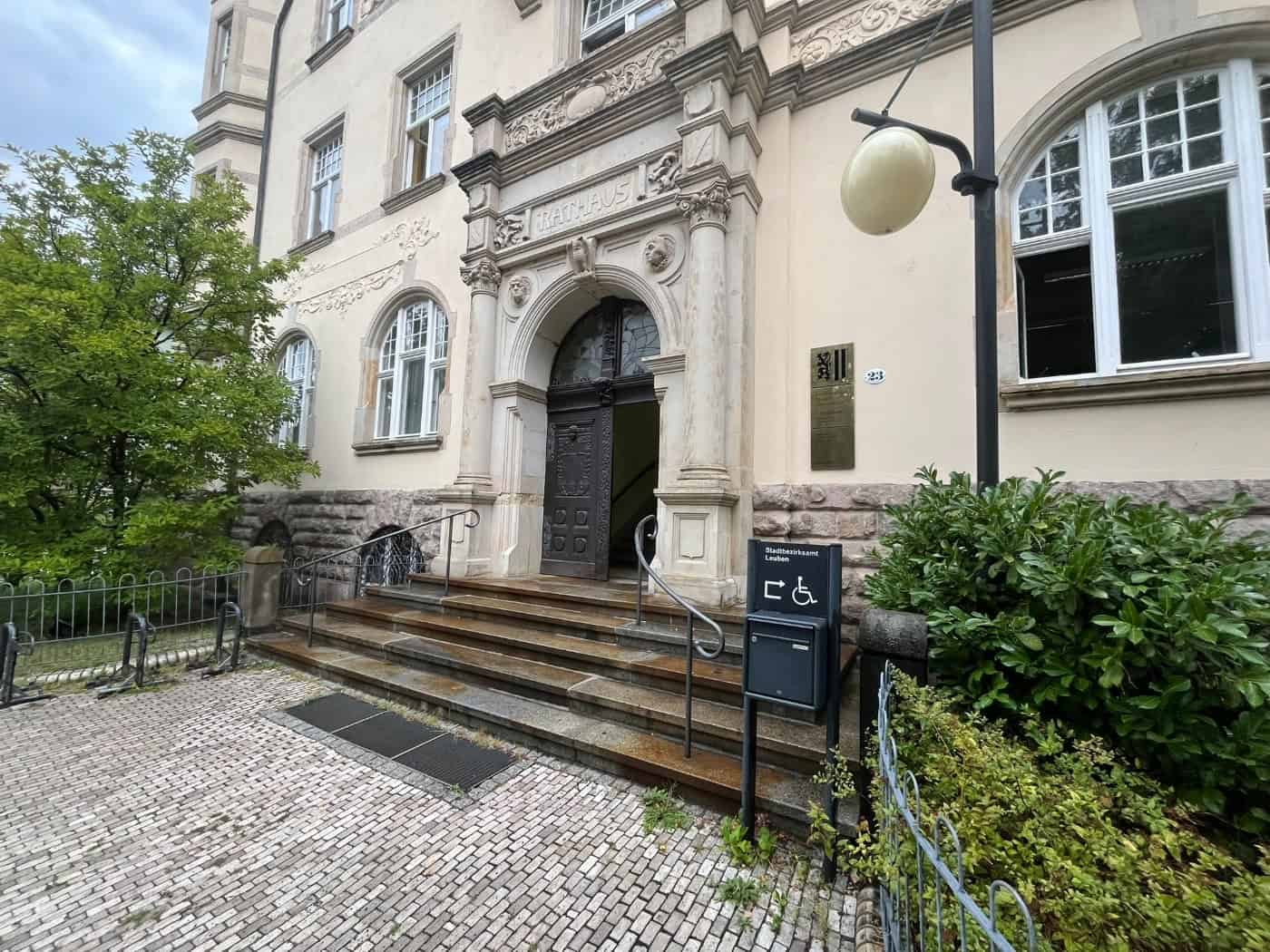 Historisches Portal des Stadtbezirksamts Leuben mit dem Schriftzug „Rathaus“, gepflastertem Vorplatz, Freitreppe und geöffnete Holztür. Rechts steht ein Hinweisschild auf den barrierefreien Zugang.