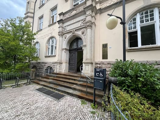 Historisches Portal des Stadtbezirksamts Leuben mit dem Schriftzug „Rathaus“, gepflastertem Vorplatz, Freitreppe und geöffnete Holztür. Rechts steht ein Hinweisschild auf den barrierefreien Zugang.
