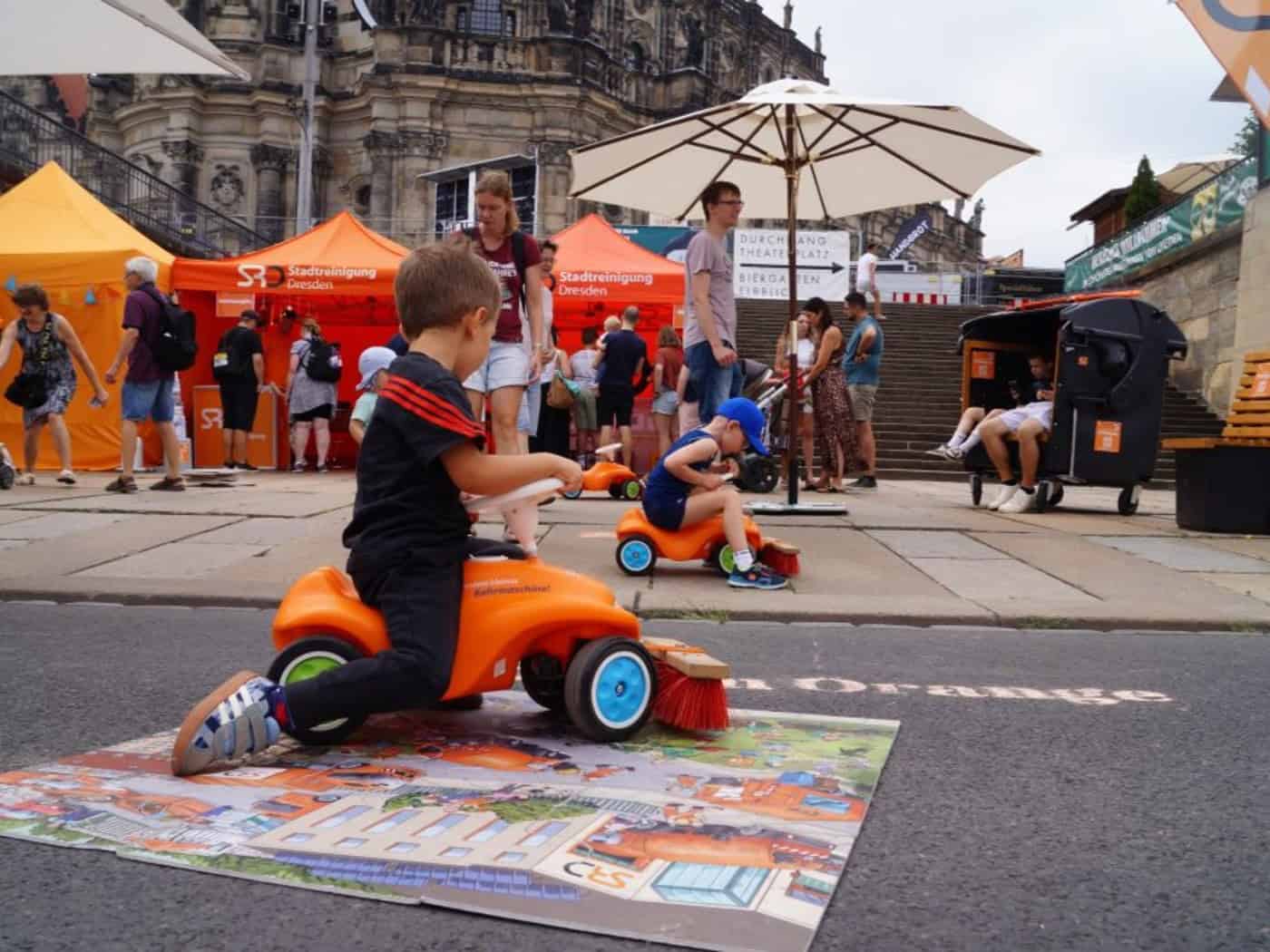 Ein kleiner Junge sitzt im Vordergrund auf einer orangefarbenen Mini-Kehrmaschine im Bobbycar-Stil. Im Hintergrund stehen orangefarbene Informationszelte der Stadtreinigung Dresden vor historischer Kulisse mit Passanten.