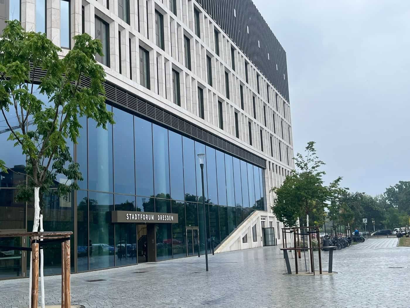 Außenansicht des modernen Gebäudes Stadtforum Dresden an der Waisenhausstraße mit großer Glasfront, hellem Stein, gepflastertem Vorplatz und jungen Bäumen unter bedecktem Himmel. 