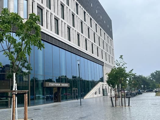 Außenansicht des modernen Gebäudes Stadtforum Dresden an der Waisenhausstraße mit großer Glasfront, hellem Stein, gepflastertem Vorplatz und jungen Bäumen unter bedecktem Himmel. 
